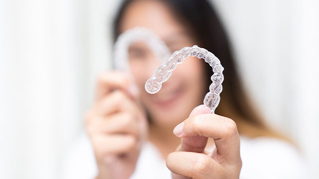 The image shows a person s hand holding up two transparent dental retainers with a blurred background.